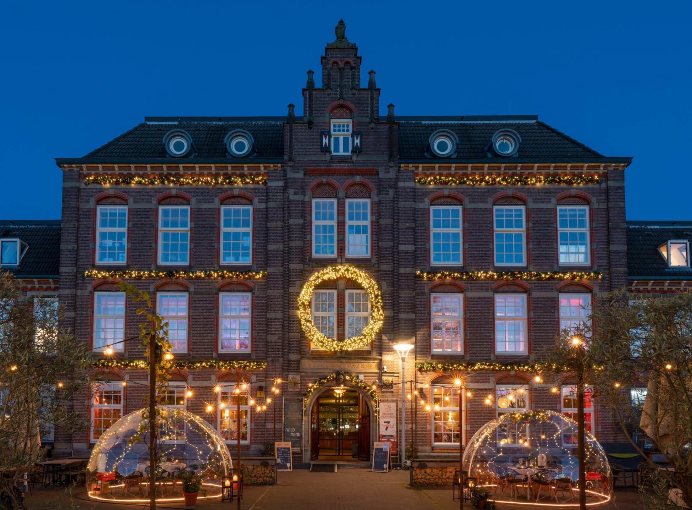 Het kazernegebouw van In de Kazerne bij schemering met de twee verlichte domes in de tuin ervoor.